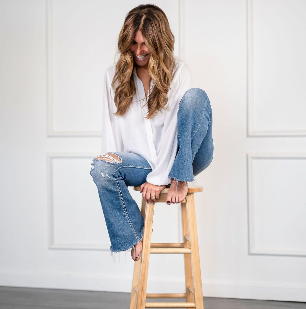 Andrea Kind sitting thoughtfully on a wooden stool, wearing a white shirt and blue jeans.